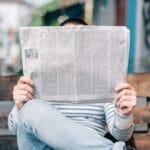 Person sitting on a bench holding a newspaper in front of their face